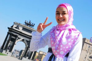 A woman in a bright pink hijab, smiling and making a peace sign.