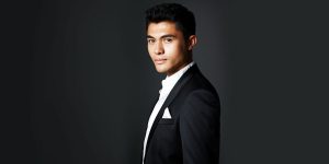 A promotional still of Henry Golding, a Eurasian actor wearing a suit.