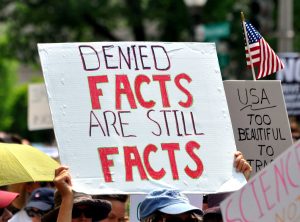 A protest sign: DENIED FACTS ARE STILL FACTS.