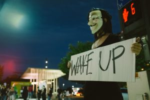 A protester in an anonymous mask carrying a sign that says WAKE UP