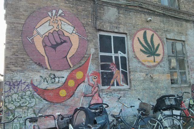 A mural on the side of a brick building in Christiana, featuring a marijuana leaf and a fist breaking a syringe.