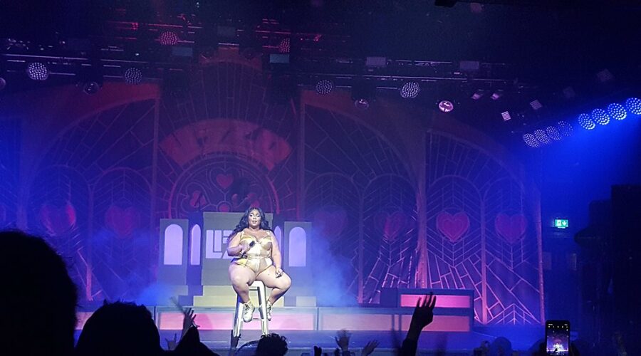 Lizzo performing in the Olympia, Dublin in 2019. She is sitting on a high stool with a microphone.